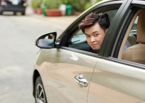 Young driver leaning out of a car window on a residential street, looking at the camera.
